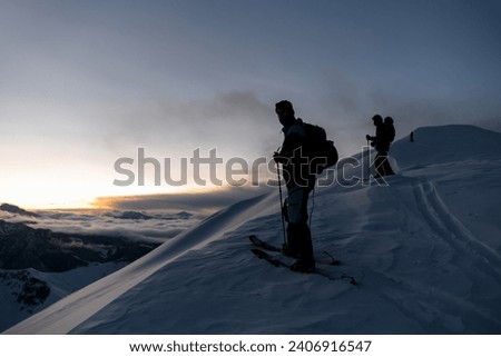Silhouettes of three skiers climbing a mountain slope against the background of lower mountains that are shrouded in fog, all placed at a distance from each other