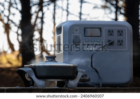 Portable Power Station heating a electric frying pan on a picnic table - steam rises - in background trees Royalty-Free Stock Photo #2406960107