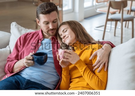 Pleased couple enjoying lazy weekend resting at home drinking coffee together on cozy sofa. Husband tender hugging wife with closed eyes. Intimacy, affectionate marrieds. Happy marriage, daydreaming Royalty-Free Stock Photo #2407006351