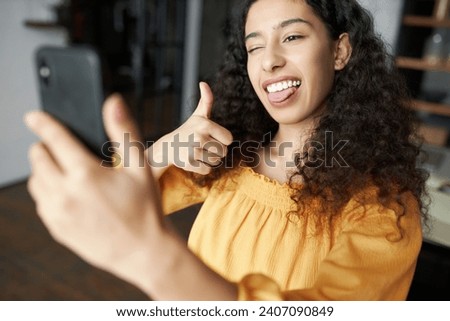 Selective focus on funny african american young female posing in front of camera of her smartphone, taking selfie, winking and showing thumb up, sticking tongue, grimacing and having fun
