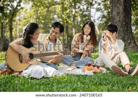 A lovely young Asian couple is playing an acoustic guitar together while enjoying a picnic with their friends in a green park on summer. Friendship concept