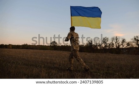 Soldier of ukrainian army running with raised blue-yellow banner on field at dusk. Young male military in uniform jogging with flag of Ukraine at meadow. Victory against russian aggression concept