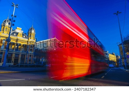 Budapest, Hungary: December 16, 2023: Long exposure shot of red bus. Restored Nyugati Train Station on the background. 