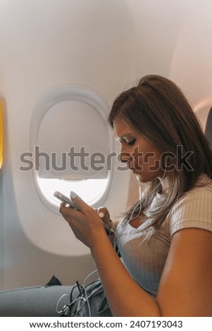 Young girl sitting on the plane looking at her cell phone with headphones on. Woman listening to music through headphones while using the airline.