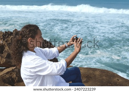 Curly haired multi ethnic pretty woman taking photo on her smart mobile phone, sitting on the rock on the Atlantic ocean beach. People and nature. Connection with nature. Recreation. Leisure activity