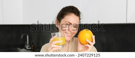 Image of good-looking healthy woman in bathrobe, drinking fresh juice, showing orange fruit, posing in kitchen.