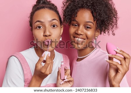 Photo of female friends apply lip gloss undergo beauty procedures at home have long nails get ready for disco party stand closely to each other isolated over pink background take care of themselves