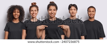 Horizontal shot of cheerful mixed race women and men dressed in black t shirts Latin woman points at t shirt for your logo isolated over white background. Happy five people stand in row in studio