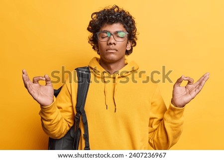 Photo of relaxed Hindu man with curly hair meditates indoor keeps eyes closed breathes deeply dressed in casual sweatshirt carries rucksack isolated over yellow background. Body language concept