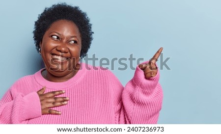 Plump dark skinned woman with curly hair points index finger on copy space for your advertisement indicates at upper right corner dressed in loose knitted jumper isolated over blue background