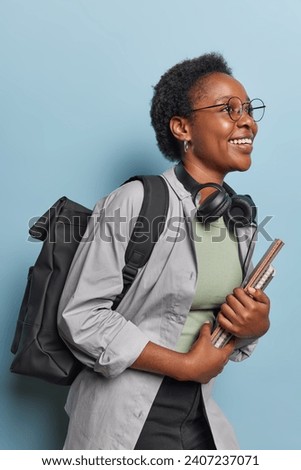 Sideways shot of cheerful millennial African girl with curly hair carries notepads wears headphones around neck carries rucksack on back concentrated somewhere has glad expression dressed casually Royalty-Free Stock Photo #2407237071