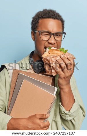 Vertical shot of handsome dark skinned man with curly hair eats sandwich as feels hungry after lectures poses with spiral notebooks wears spectacles and casual shirt isolated over blue background Royalty-Free Stock Photo #2407237137