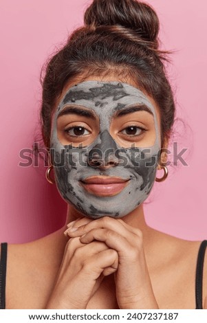 Vertical image of lovely dark haired woman keeps hands under chin concentrated at camera applies facial clay mask for skin treatment undergoes beauty procedures isolated over pink background