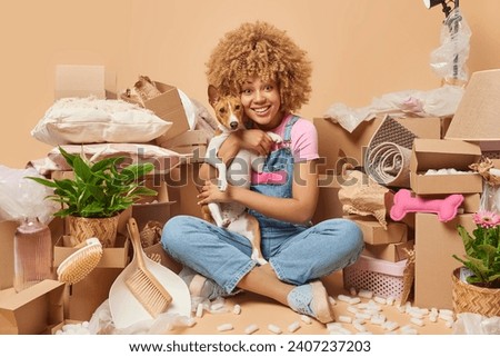 Young beautiful curly haired woman sits crossed legs with pet surrounded by carton boxes full of property going to relocate to other apartment isolated over brown background. Moving Day concept