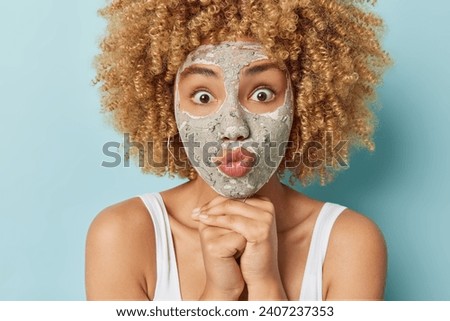 Close up shot of curly haired woman keeps hands under chin has lips rounded takes on her skincare routine adding clay mask for radiant look wears white t shirt isolated over blue background.