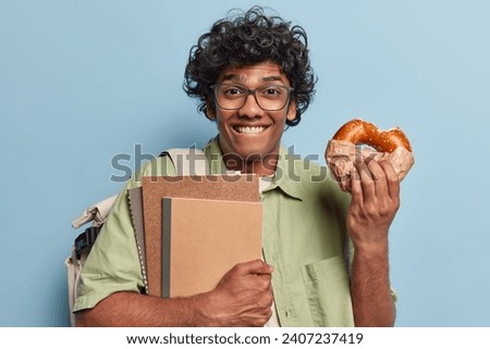 Horizontal shot of cheerful Hindu man with curly hair holds notepads and delicious bun wears transparent eyeglasses and shirt carries rucksack isolated over blue background being hungry after studying