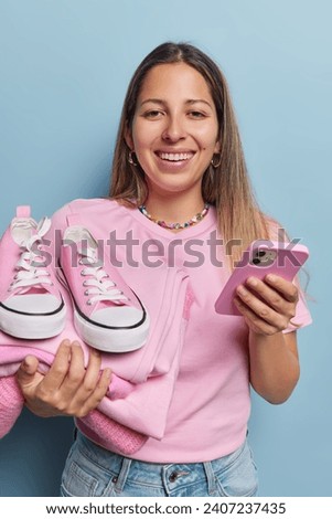 Vertical shot of cheerful long haired woman smiles broadly holds smartphone in one hand pile of ironed washed clothes and sneakers in other sells second hand items online isolated over blue background Royalty-Free Stock Photo #2407237435