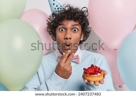 Photo of shocked Hindu man with dark curly hair celebrates special occasion holds small strawberry cupcake with burning candle surrounded by inflated colorful balloons reacts to something shocking