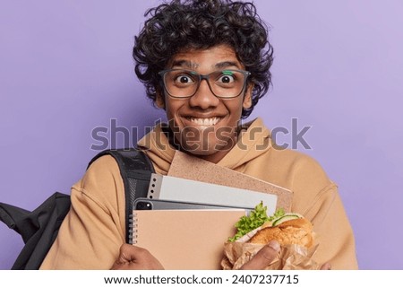 People positive emotions concept. Indoor photo of young cheerful smiling Hindu male student standing in centre isolated on purple background holding books tablet notebooks and tasty sandwich