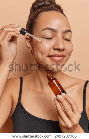 Beauty and skin care concept. Young woman applies vitamin c or essential oil keeps eyes closed dressed in black t shirt uses natural organic cosmetic product for skin regeneration poses indoor