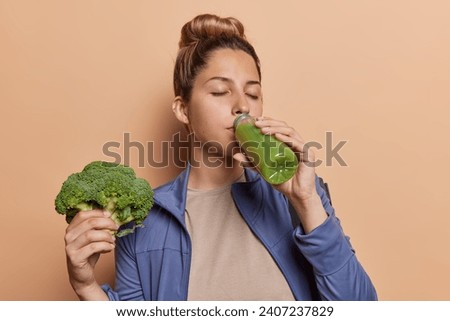 Sporty concept. Indoor photo of young satisfied glad fit European woman wearing tracksuit standing in centre on beige background holding brocolli drinking green smoothie keeping healthy lifestyle Royalty-Free Stock Photo #2407237829