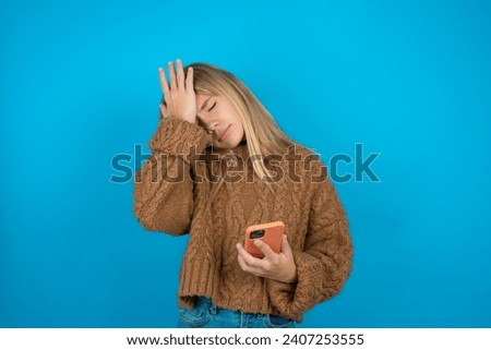 Upset depressed Beautiful kid girl wearing brown knitted sweater makes face palm as forgot about something important holds mobile phone expresses sorrow and regret blames Royalty-Free Stock Photo #2407253555