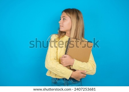 Beautiful kid girl wearing yellow sweater holding notebook arms crossed look empty space billboard