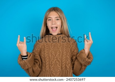 Beautiful kid girl wearing brown knitted sweater makes rock n roll sign looks self confident and cheerful enjoys cool music at party. Body language concept.