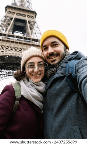 Vertical photo of young adult european couple taking selfie portrait with phone in front of Eiffel Tower in Paris city, France. Travel and vacation concept.