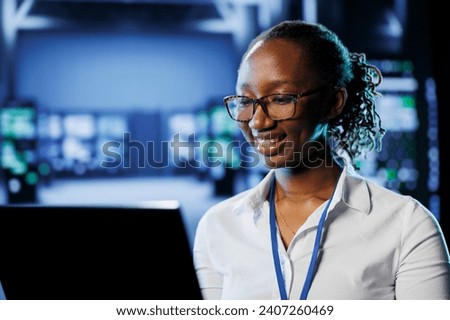 Joyful admin in high tech workplace uses laptop to prevent system overload. Competent expert between server racks ensuring enough network bandwidth for smooth operations Royalty-Free Stock Photo #2407260469