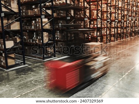Blur of a motorized stock picker between aisles of cardboard boxes on pallets stacked on large racks in a large distibution warehouse.