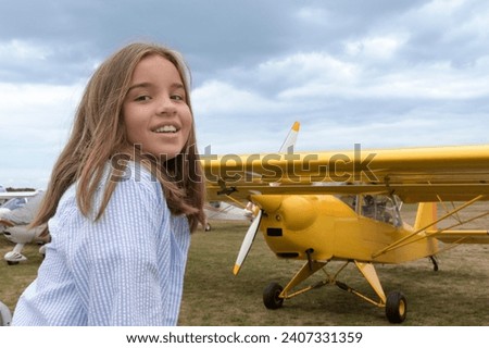 Smiling teenage girl in front of a vintage yellow plane on the airfield Royalty-Free Stock Photo #2407331359