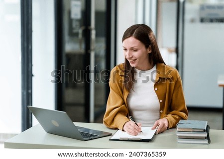 Caucasian university woman working on a project, with a laptop, clipboard in hand, and books on the table.