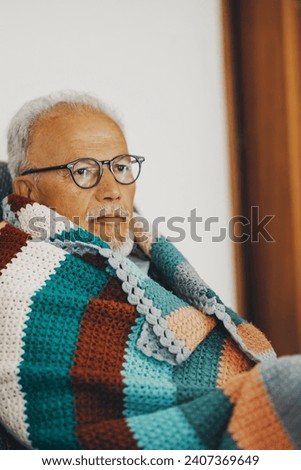One old senior man sitting on the sofa at home with colorful wool cover for low temperature and cold flu symptoms. Energy gas crisis concept. Savings heat indoor. Concept of people in winter