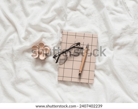 Stylish women's accessories on white sheets in bed - a notebook, glasses, a flower hairpin, a pen. Cozy morning concept