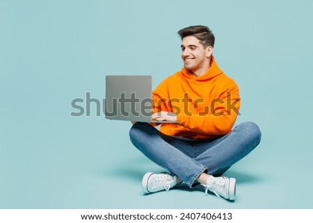 Full body young IT man he wearing orange hoody casual clothes sitting hold use work on laptop pc computer isolated on plain pastel light blue cyan color background studio portrait. Lifestyle concept Royalty-Free Stock Photo #2407406413