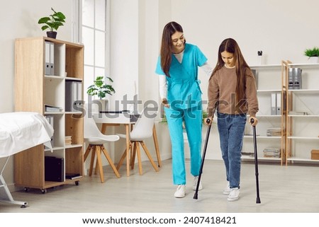 Woman doctor or nurse in a medical scrubs uniform helps a little kid child patient girl walk with crutches at the clinic during a rehabilitation course after her leg injury Royalty-Free Stock Photo #2407418421