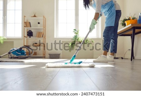 Housewife or professional cleaner mopping the floor at home. Crop young woman in an apron and gloves washing light gray wooden or laminate flooring in the living room. Housework, cleaning concept Royalty-Free Stock Photo #2407473151