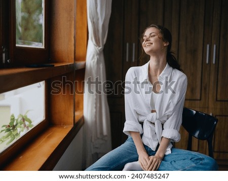 Woman sitting at home by a wooden window with a smile on a chair in homemade comfortable clothes and looking at the landscape, spring mood, women's day, rest on the weekend. Royalty-Free Stock Photo #2407485247