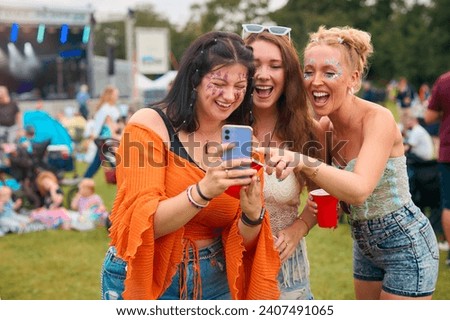 Three Female Friends Wearing Glitter Looking At Mobile Phone At Summer Music Festival Holding Drinks