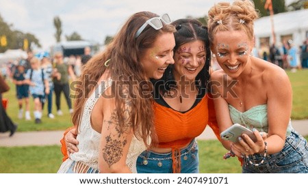 Three Female Friends Wearing Glitter Looking At Mobile Phone At Summer Music Festival