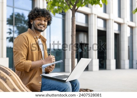 Portrait of a smiling fashionable Indian man studying and working remotely. Sitting on a bench on the street with a laptop and headphones, holding a notebook and looking at the camera.
