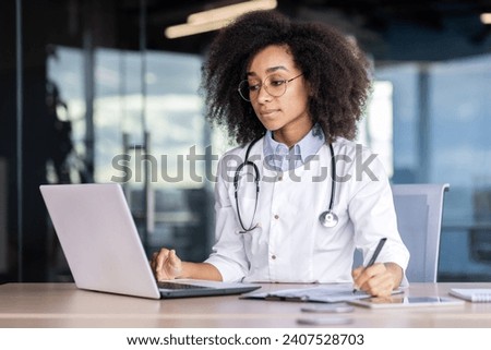 Concentrated young African American female doctor, student practitioner studying and working online on laptop, sitting at desk in office and taking notes. Royalty-Free Stock Photo #2407528703