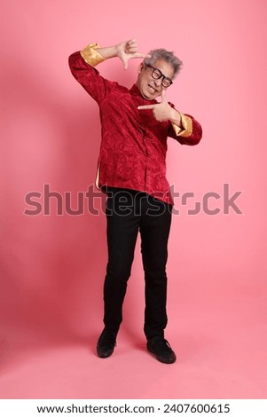 Happy Chinese new year. Asian Chinese energetic senior man wearing red traditional cheongsam qipao or changshan dress with gesture of selfie isolated on pink background.