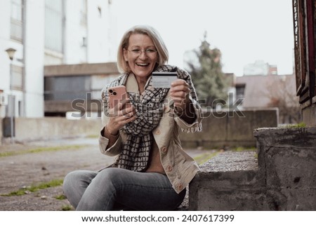 mature blonde woman sitting outside in a residential area of ​​the city and shopping with a credit card online