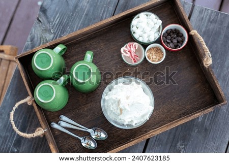 Hot chocolate bar concept. 3green mugs with bowl of whipped cream and toppings in different holiday bowls; mini marshmallows, chocolate chips, miniature candy canes, toffee pieces, on a wood tray. Royalty-Free Stock Photo #2407623185