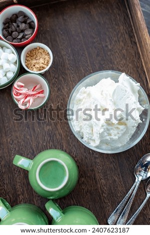 Hot chocolate bar concept. 3green mugs with bowl of whipped cream and toppings in different holiday bowls; mini marshmallows, chocolate chips, miniature candy canes, toffee pieces, on a wood tray. Royalty-Free Stock Photo #2407623187