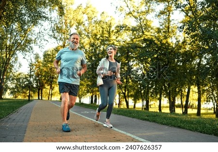 Sport outdoors. Positive man and woman with grey hair cheerfully smiling and talking while jogging together on fresh air. Caucasian family of two in sportswear and sneakers training at summer park.