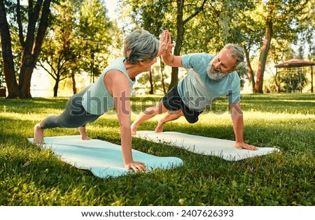Teamwork during outdoors activity. Strong elderly couple in sportswear standing in plank position, giving high five, looking at each other and smiling. Concept of healthy retirement and active hobby. Royalty-Free Stock Photo #2407626393