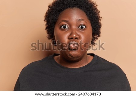 Photo of chubby African woman holds breath keeps lips rounded and stares bugged eyes at camera with funny grimace wears black t shirt isolated over brown background. Human facial expressions